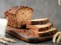 Chleb proteinowy z ziarnami słonecznika i dyni, pokrojony na kromki, obok szklanka mleka i kłosy zboża.