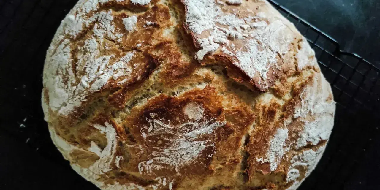 Chleb rustykalny z chrupiącą skórką i puszystym wnętrzem, posypany mąką, leży na czarnej kratce.
