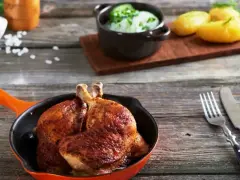 Złociste udka z airfryer w pomarańczowej patelni, obok ziemniaczki i miseczka z zielonym sosem.
