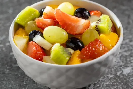 Kolorowa sałatka owocowa składniki: truskawki, kiwi, winogrona, mango i czarne porzeczki w białej miseczce.