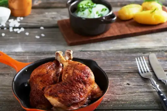 Złociste udka z airfryer w pomarańczowej patelni, obok ziemniaczki i miseczka z zielonym sosem.