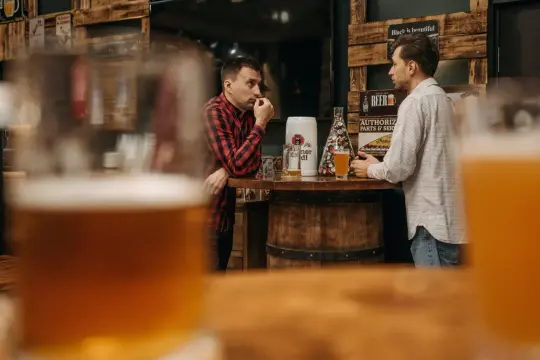 Dwóch mężczyzn rozmawia przy barze z piwem, w tle dekoracje związane z piwem.