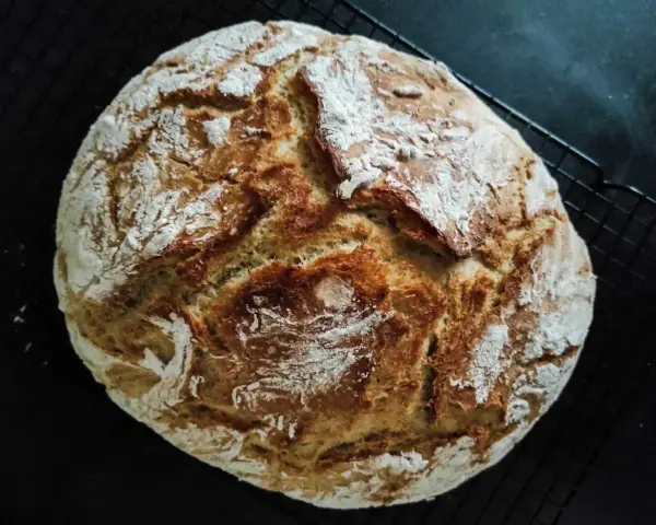 Chleb rustykalny z chrupiącą skórką i puszystym wnętrzem, posypany mąką, leży na czarnej kratce.